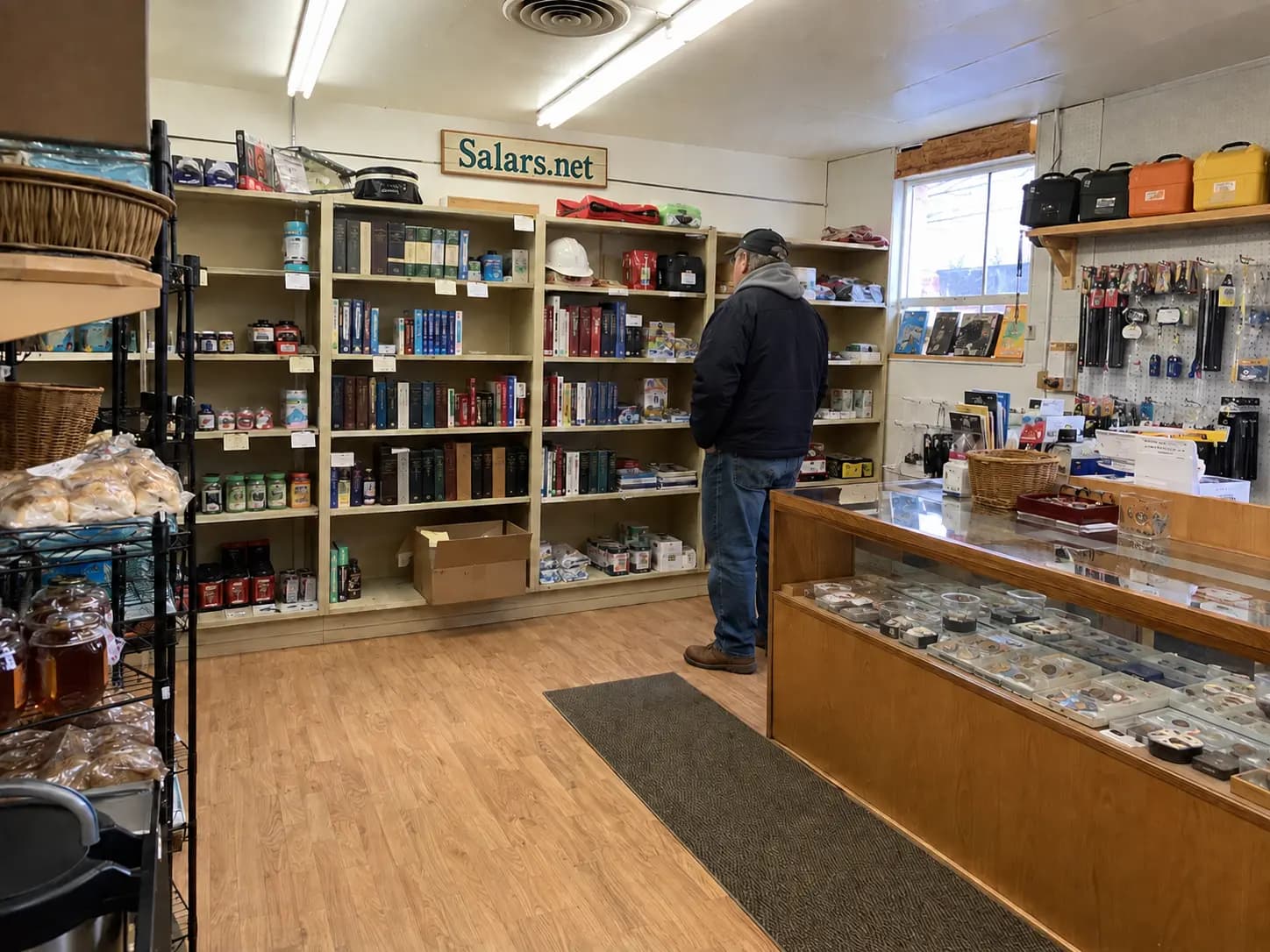 Representative interior view of a small Salars.net shop with coins, books, practical gear, local food, and a shopper browsing