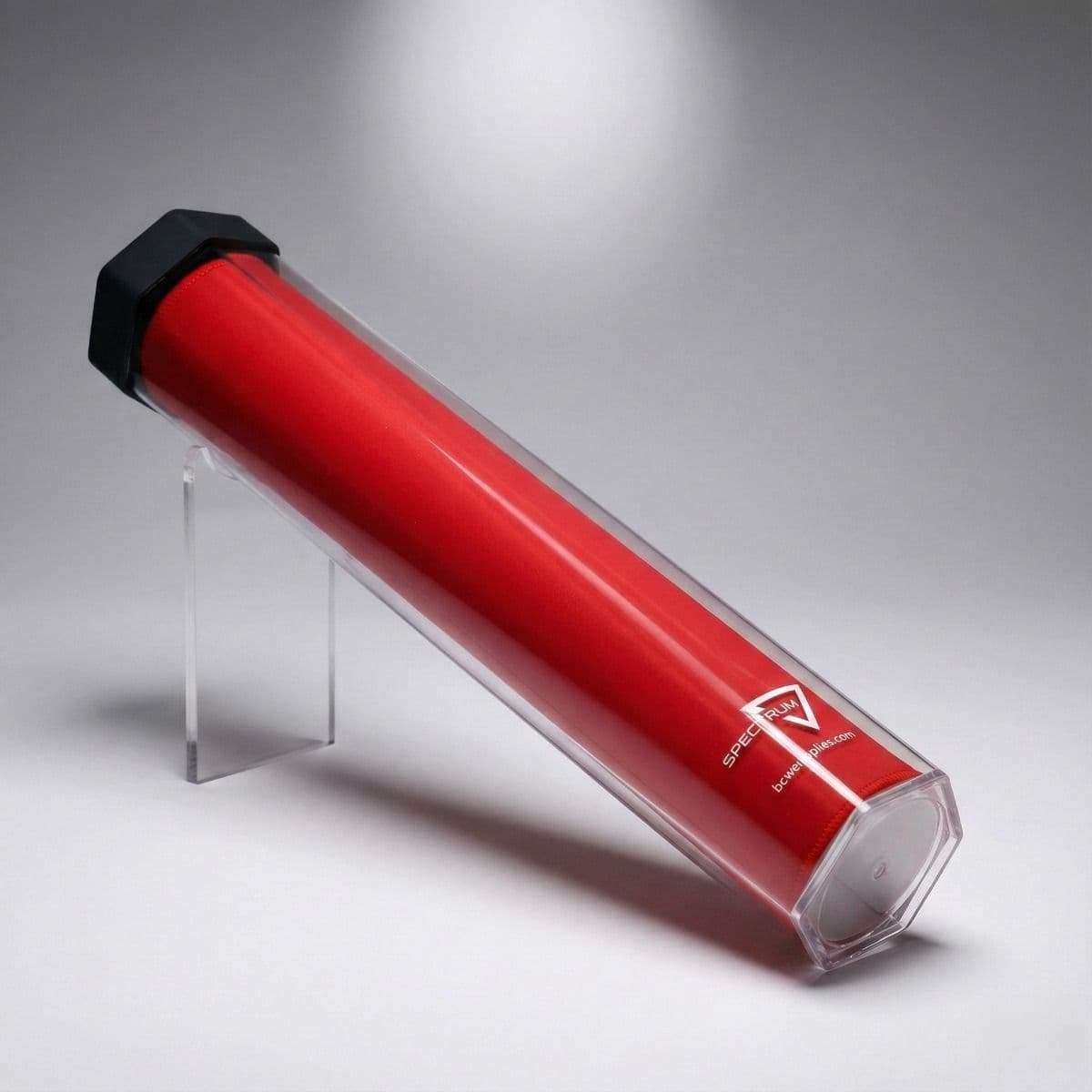 A red playmat is stored inside a clear hexagon-shaped tube with a black cap, displayed on a stand to showcase protective storage and sleek design.
