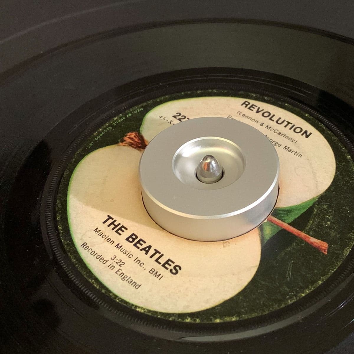 BCW 7 Inch Record Player Adapter - Classic in use with a 7-inch vinyl record of 'Revolution' by The Beatles. The silver aluminum adapter fits securely in the center hole of the record, providing stability on the turntable for smooth playback. The iconic A