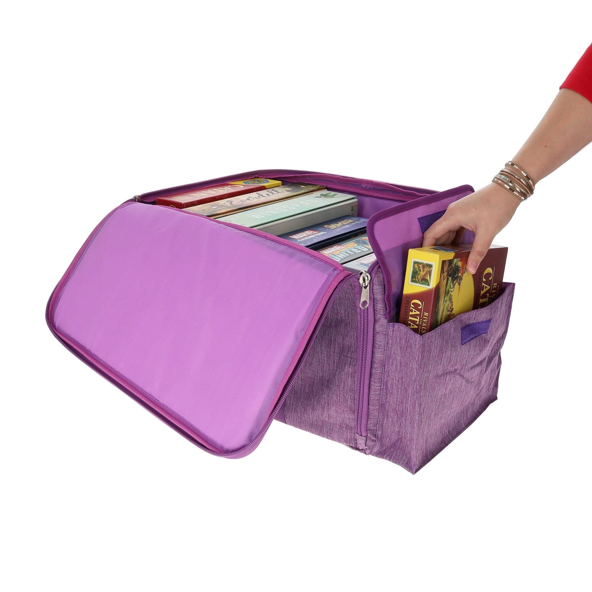 the BCW Board Game Bag in purple, with a person's hand placing the "Catan" game into one of the bag's expandable side pockets. The bag is partially unzipped to show the main compartment.