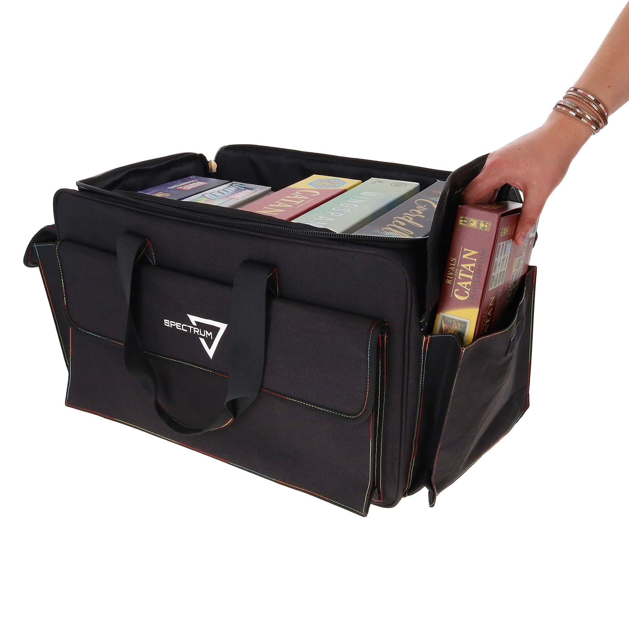the BCW Board Game Bag in black, with a person's hand placing the "Catan" game into one of the bag's expandable side pockets. The bag is partially unzipped to show the main compartment.