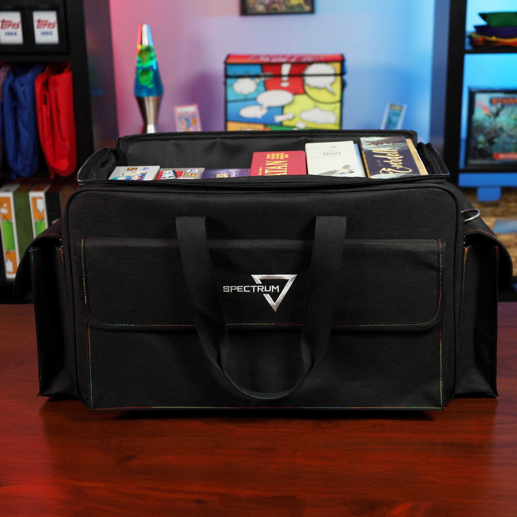the BCW Board Game Bag in black, positioned on a wooden tabletop in a game room setting. The bag's top is partially open, displaying several board games inside.