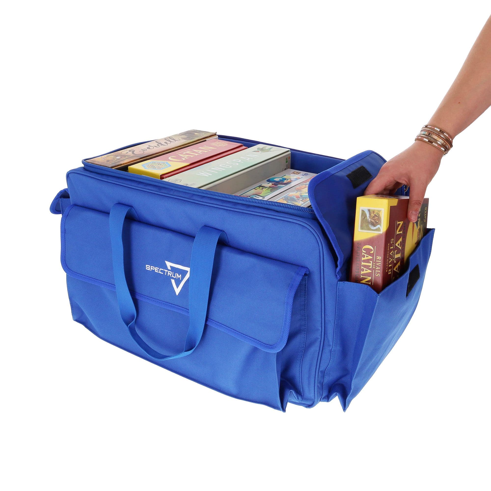 the BCW Board Game Bag in blue, with a person's hand placing the "Catan" game into one of the bag's expandable side pockets. The bag is partially unzipped to show the main compartment.