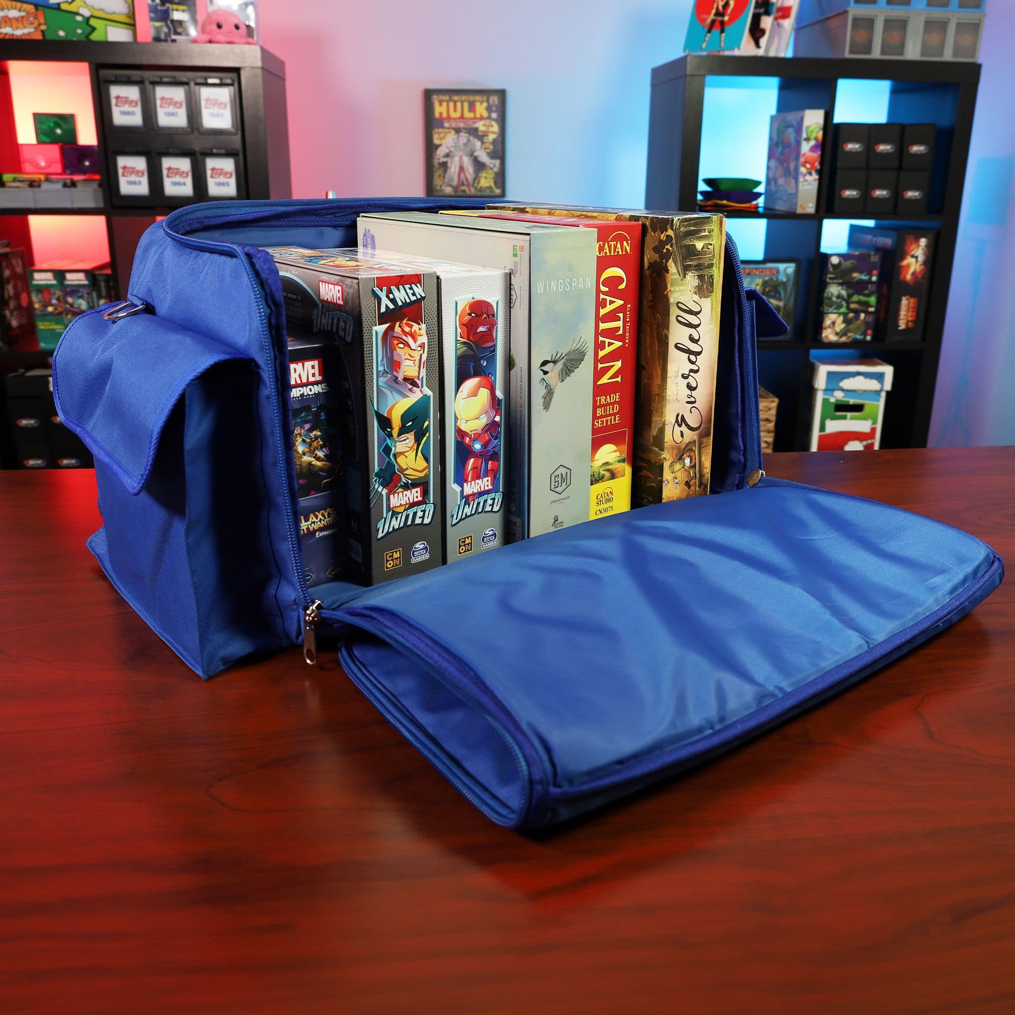 the BCW Board Game Bag in blue, placed on a wooden tabletop with the top unzipped and laid open to reveal its contents. Several board games are neatly organized inside the bag.