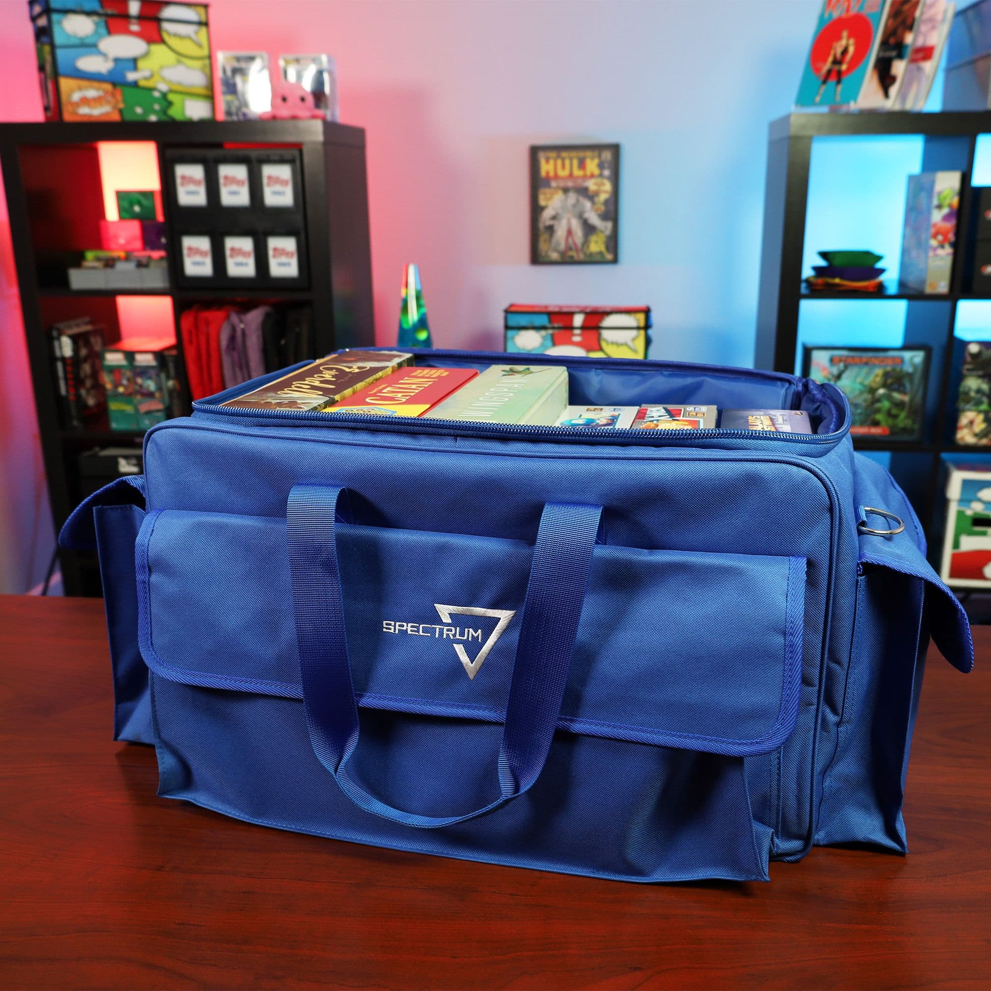 the BCW Board Game Bag in blue, positioned on a wooden tabletop in a game room setting. The bag's top is partially open, displaying several board games inside.
