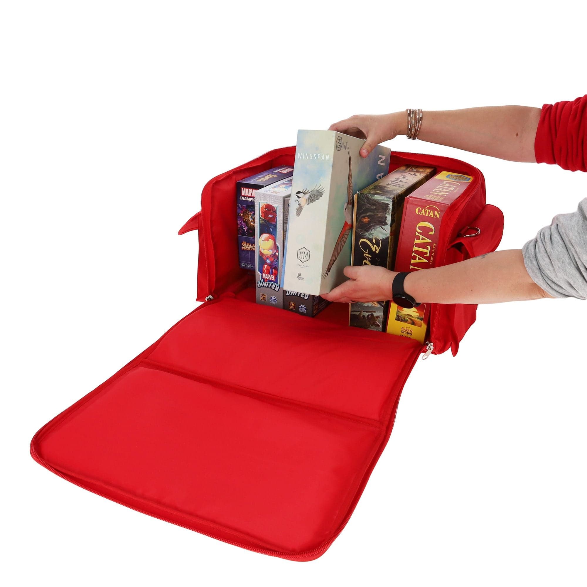 red BCW Board Game Bag being used to store several board games. The bag's top is unzipped and laid open, revealing the interior space, which can accommodate approximately six to eight standard-sized board games. A person is pulling a board game out of it