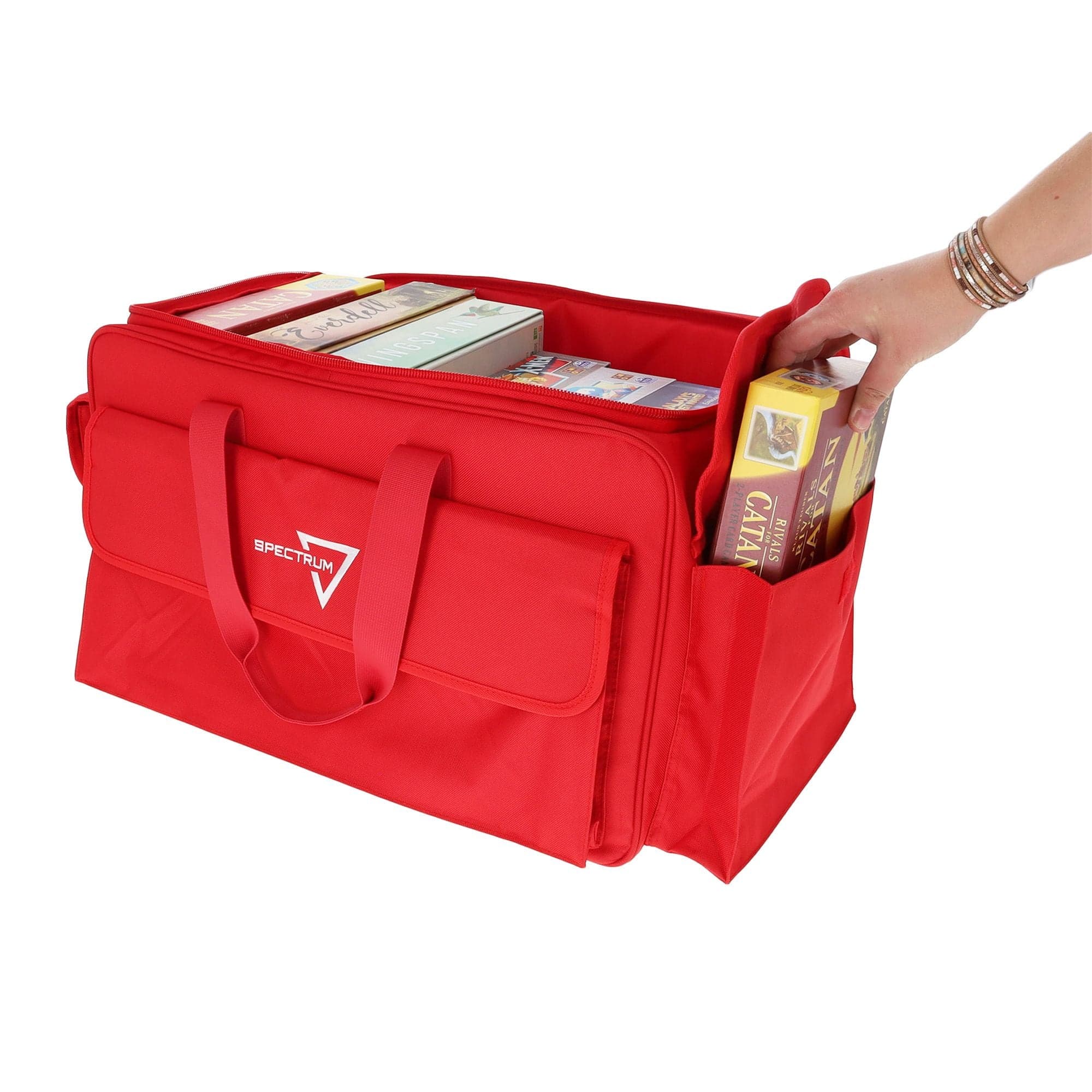 the BCW Board Game Bag in red, with a person's hand placing the "Catan" game into one of the bag's expandable side pockets. The bag is partially unzipped to show the main compartment.