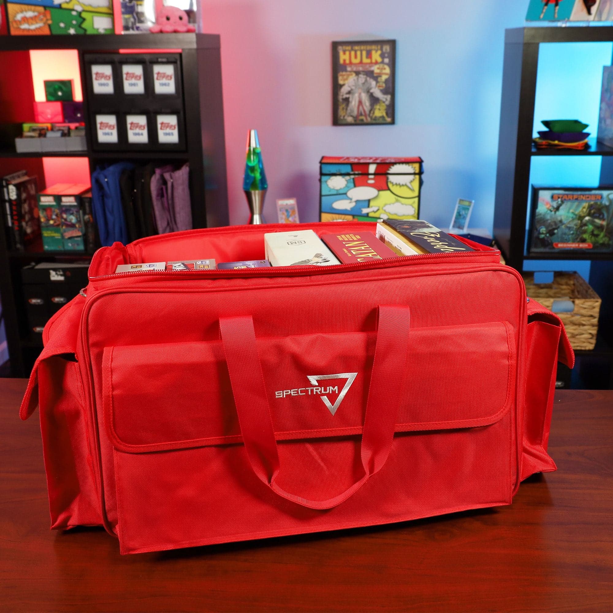 the BCW Board Game Bag in red, positioned on a wooden tabletop in a game room setting. The bag's top is partially open, displaying several board games inside.