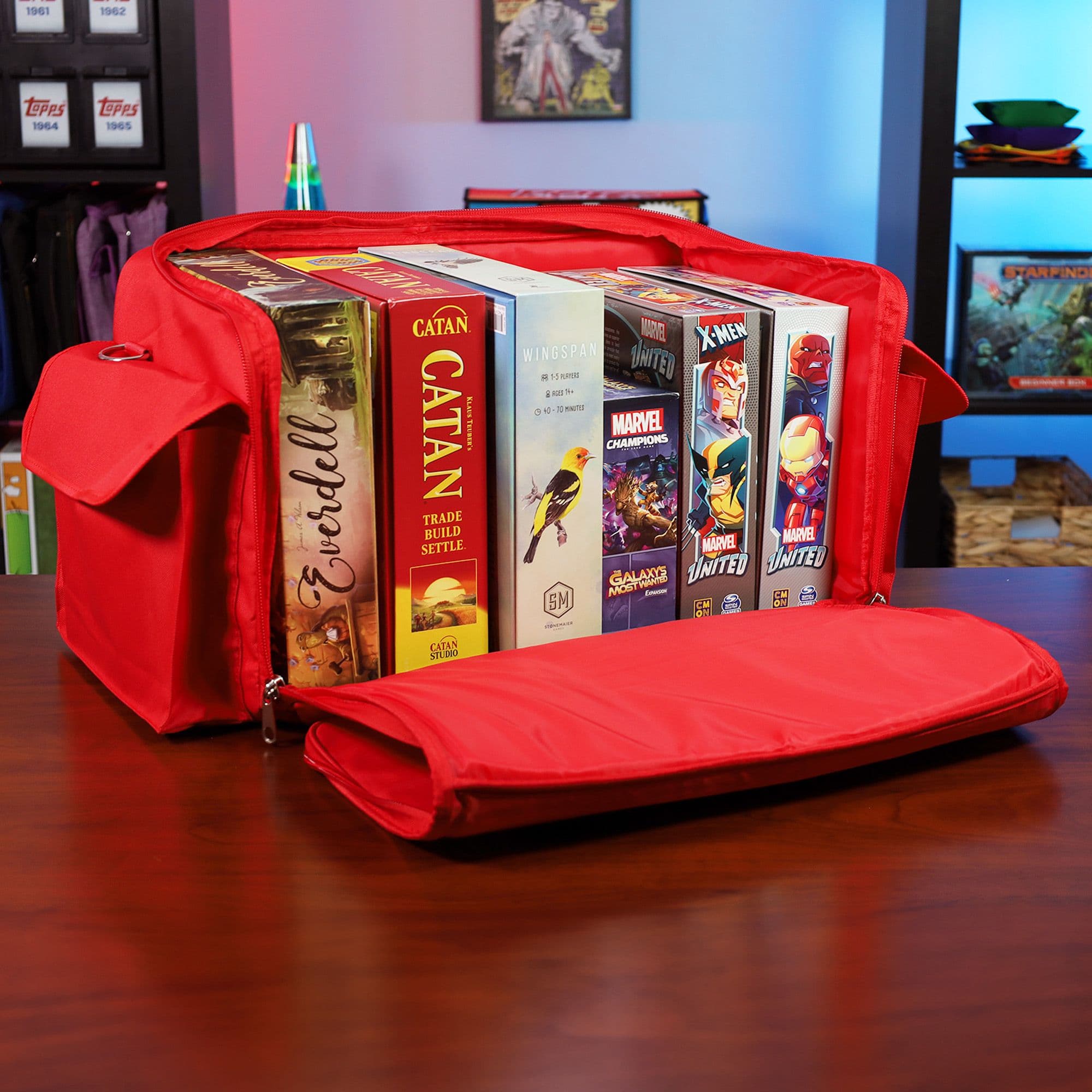 the BCW Board Game Bag in red, placed on a wooden tabletop with the top unzipped and laid open to reveal its contents. Several board games are neatly organized inside the bag.