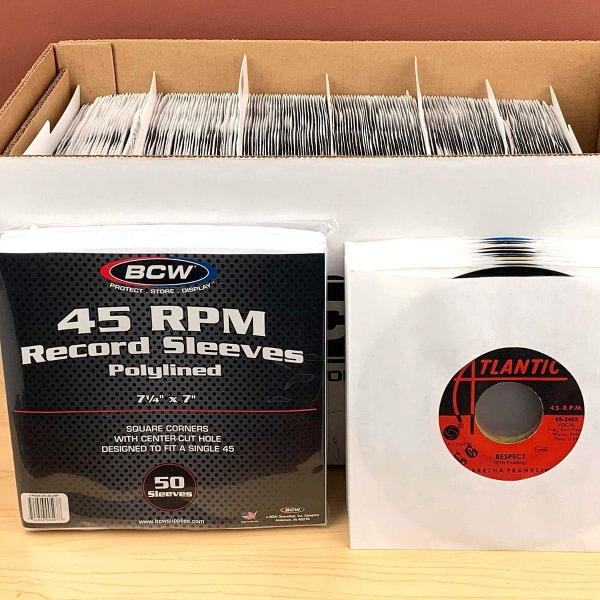 Package of BCW Paper Record Sleeves 45 RPM - Polylined - SQ Corners - With Hole next to stack of records in sleeves. These are in front of a box of records.