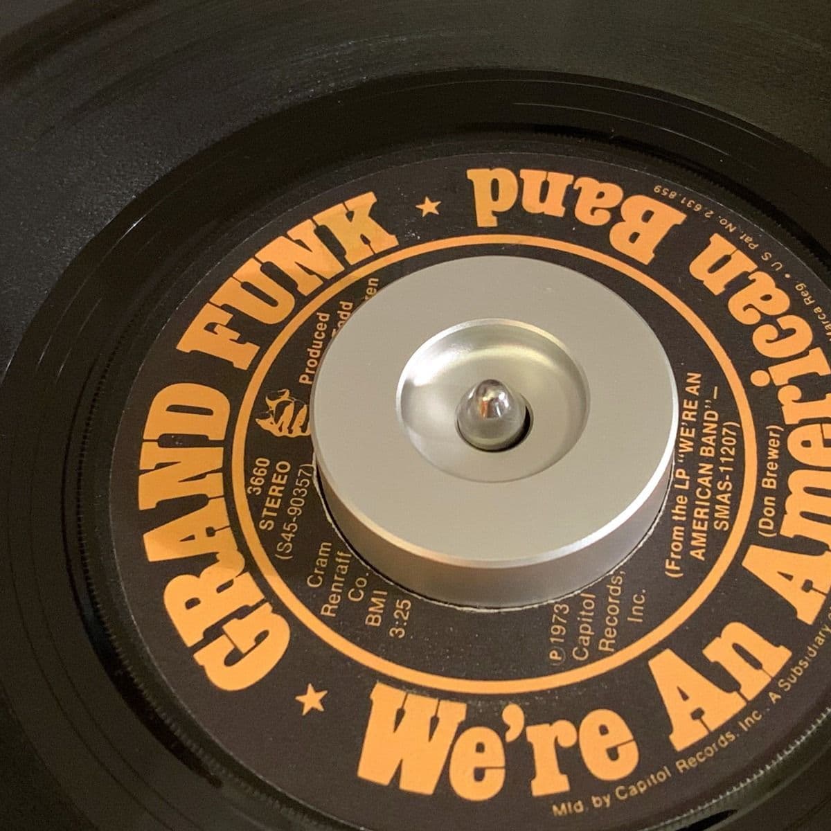 BCW 7 Inch Record Player Adapter - Classic in use with a 7-inch vinyl record of 'We're An American Band' by Grand Funk. The silver aluminum adapter fits snugly in the center hole of the record, providing stability on the turntable for smooth playback.
